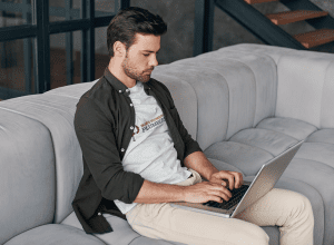 A man in his late 20s wearing a white ‘Bob’s Plumbing’ T-shirt and dark open shirt, sitting on a light grey couch and typing on a laptop, illustrating the concept of does online marketing work for small businesses.