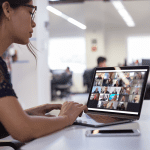 A woman working on a laptop in an open office while attending a Zoom meeting with members of an online BNI chapter shown on the screen.