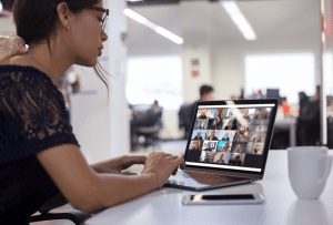 A woman working on a laptop in an open office while attending a Zoom meeting with members of an online BNI chapter shown on the screen.