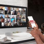 A person seated at a desk with a phone showing the BNI Online logo and a computer screen full of Zoom participant tiles, capturing a busy online BNI chapter meeting.