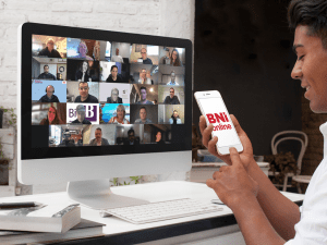 A person seated at a desk with a phone showing the BNI Online logo and a computer screen full of Zoom participant tiles, capturing a busy online BNI chapter meeting.